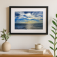 Framed artwork of a blue sky and ocean on a wall above a wooden desk with decor items.